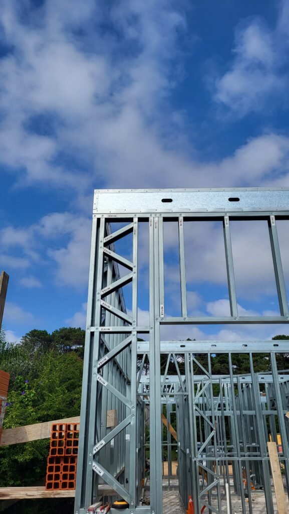 Estructura de entramado de acero galvanizado ligero (Steel Frame) en una obra de construcción al aire libre bajo un cielo azul en el Mediterráneo (Costa Blanca). Detalle de perfiles metálicos, conexiones y travesaños diagonales. Al fondo se ve vegetación verde y en primer plano unos ladrillos de arcilla tradicionales.